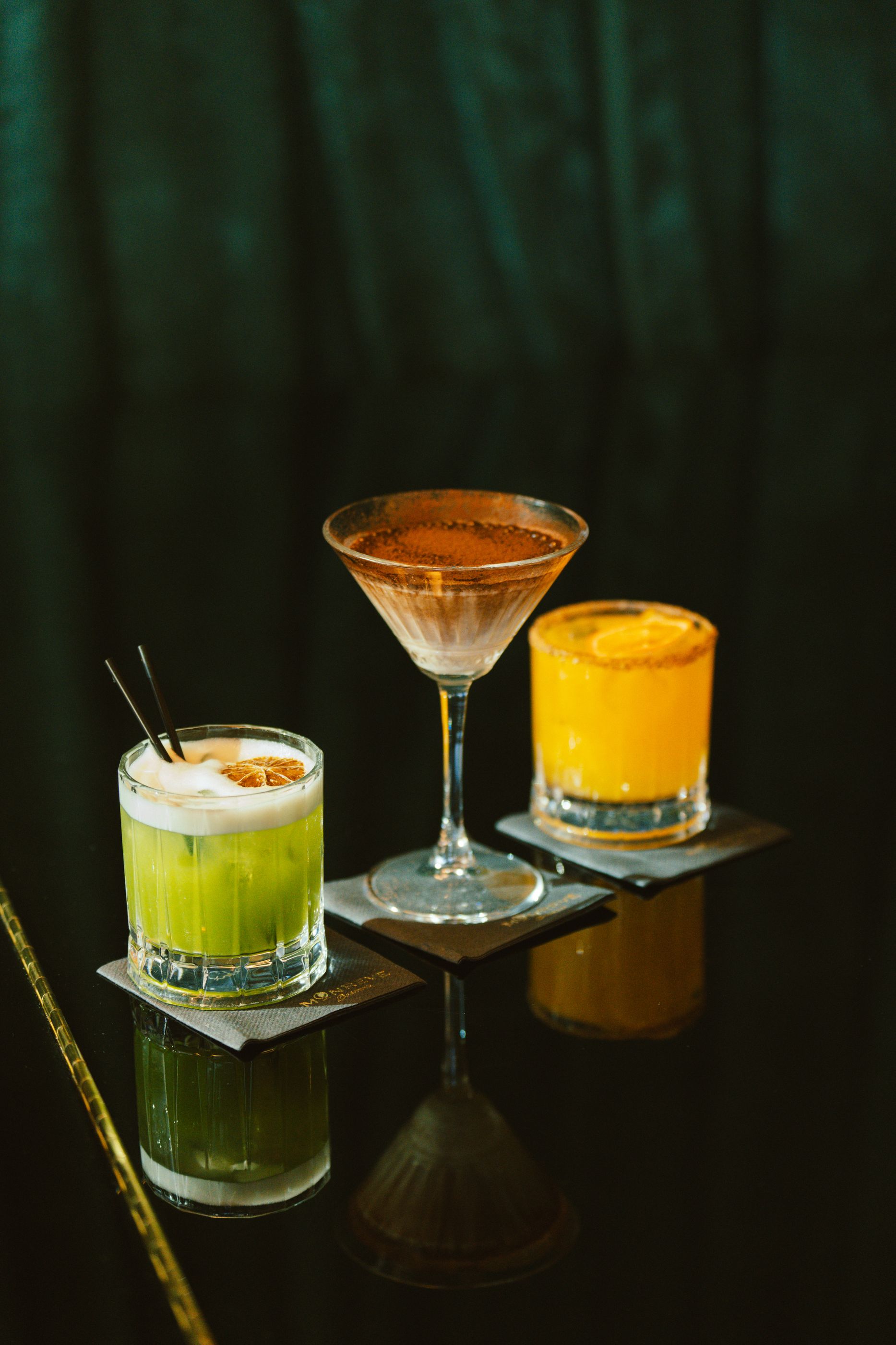 Three colourful cocktails in different glasses on a dark reflective bar counter