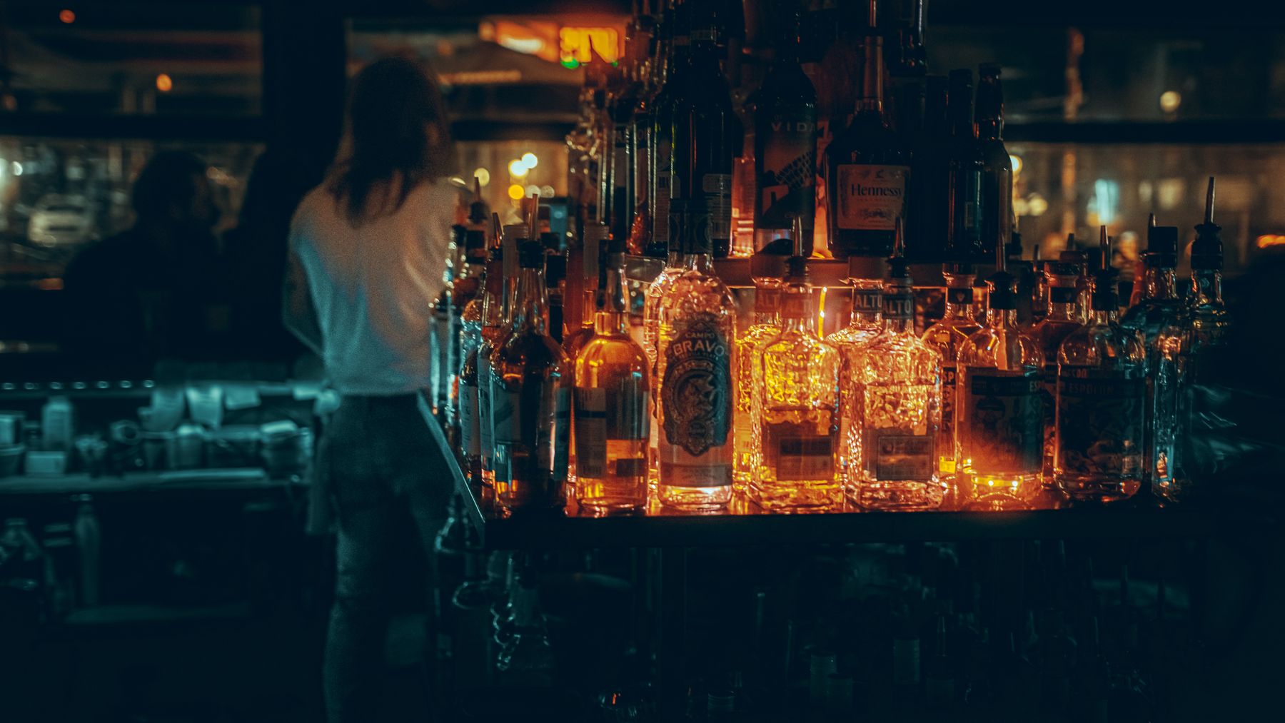 Backlit shelves of spirits bottles glow behind a bartender in a dark, atmospheric bar