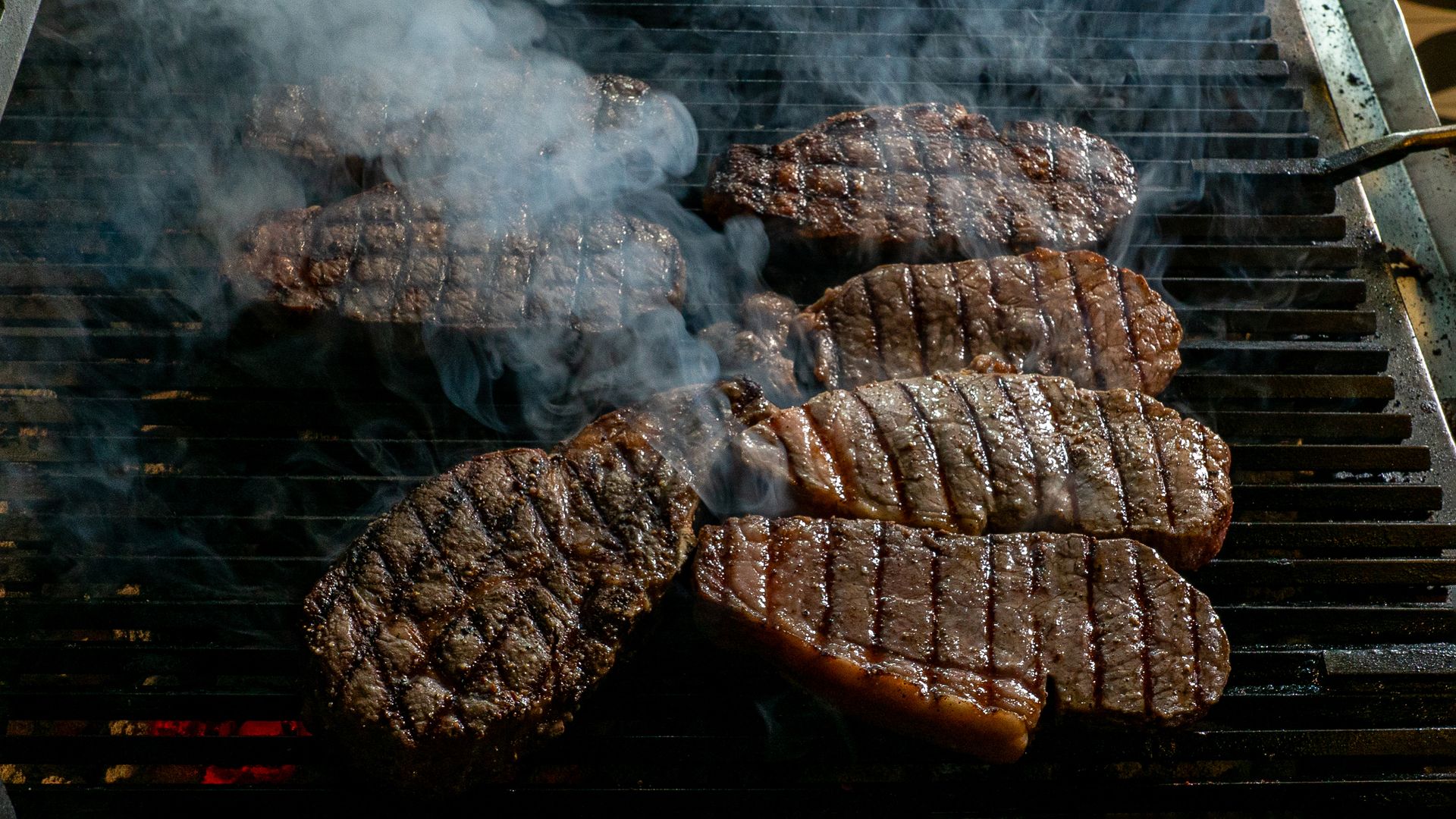 Several beef steaks searing on a charcoal grill with smoke rising