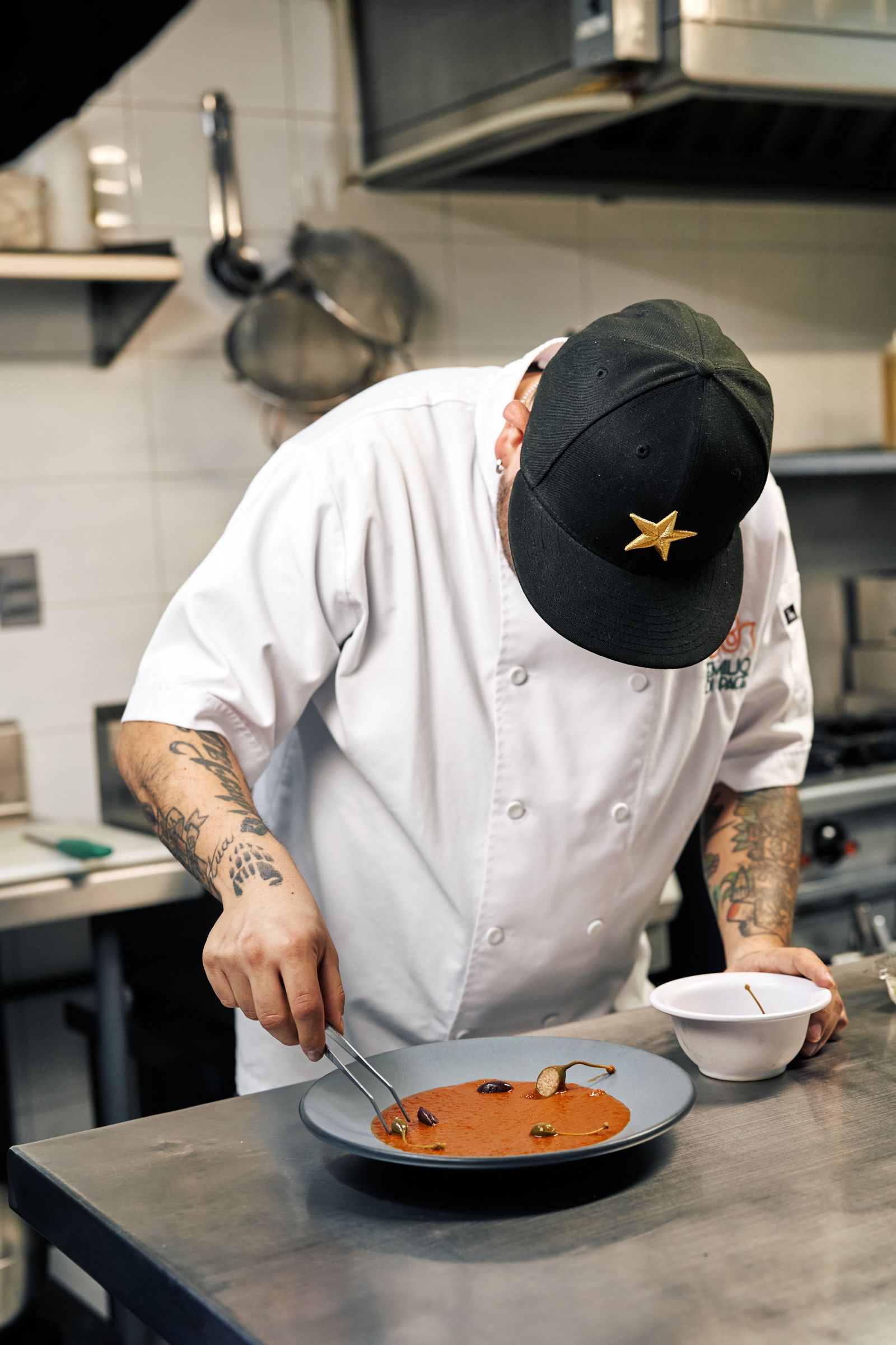 A chef finishing a plate of tomato broth with olives and capers in a professional kitchen
