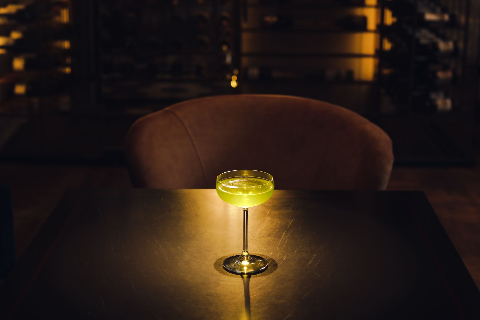 A pale green cocktail in a coupe glass sits lit on a dark bar table