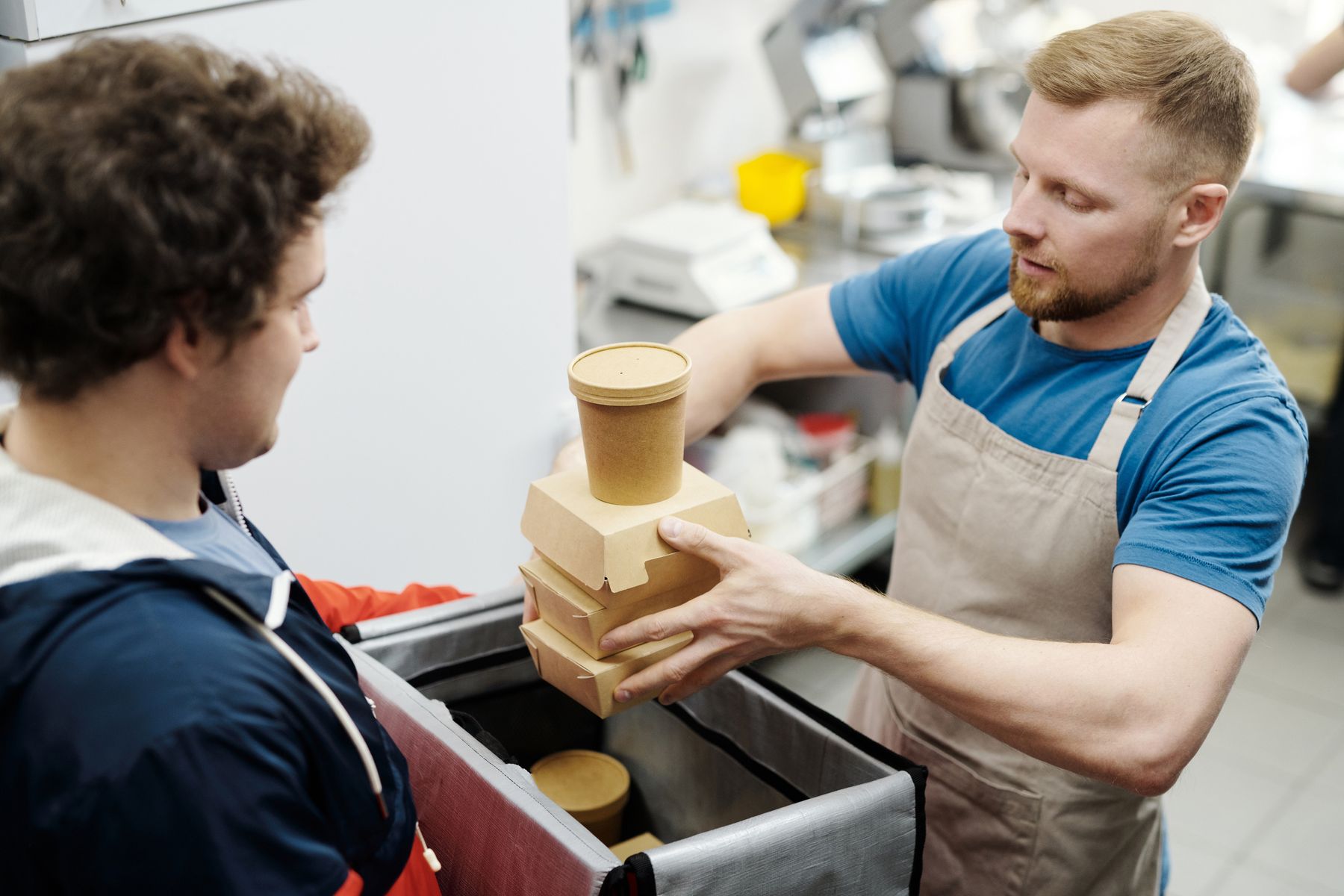 A kitchen worker in an apron hands stacked takeout containers to a delivery courier