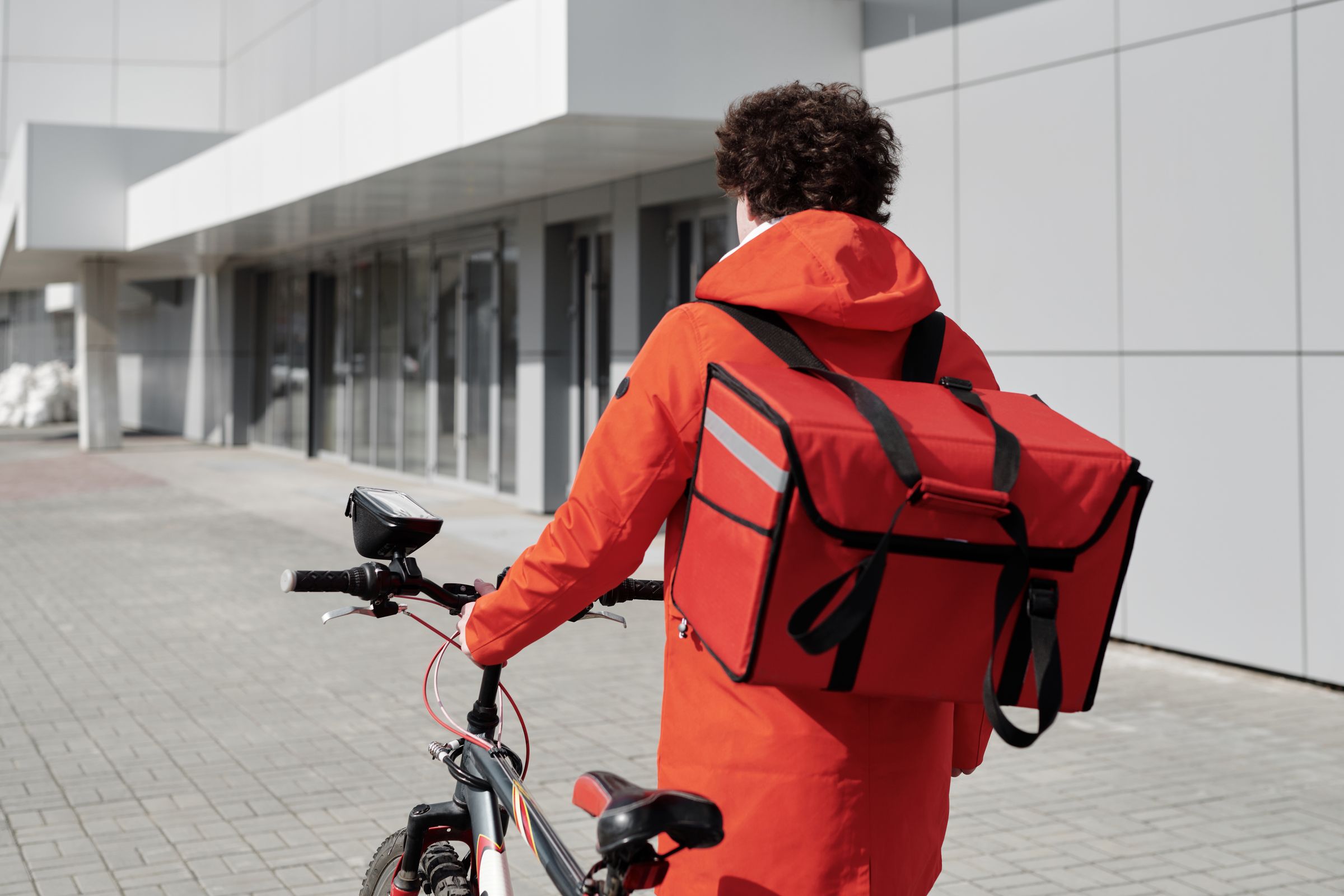 Food delivery courier in a red jacket walking a bicycle with a thermal bag in a city street