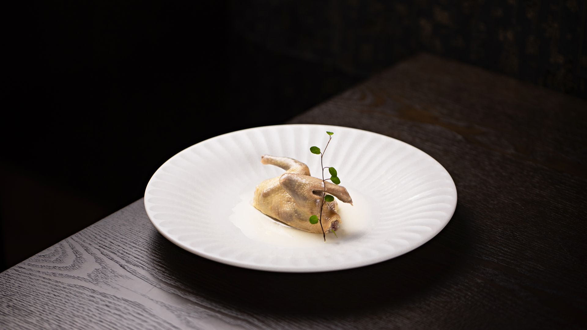 Whole roasted quail on a white fluted plate in a dark dining room