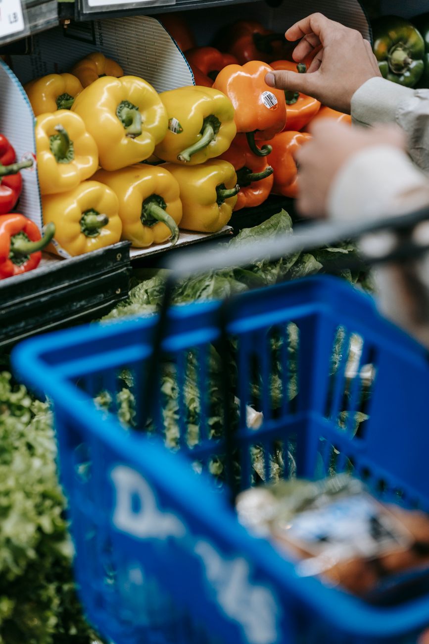 Main d'une cliente choisissant des poivrons sur une étagère de supermarché au-dessus d'un panier plein