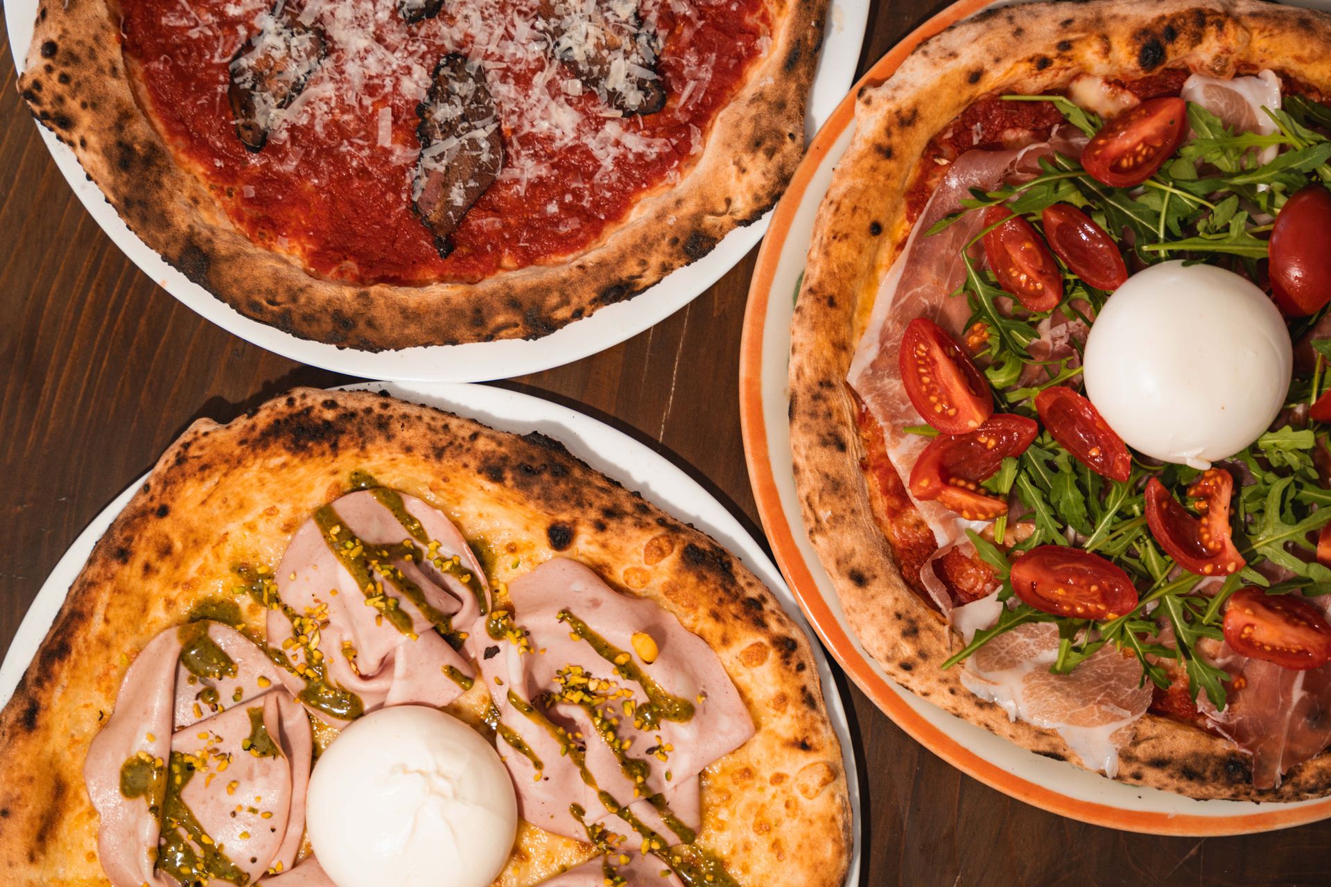 Three Neapolitan pizzas with burrata, mortadella and prosciutto on a wooden table