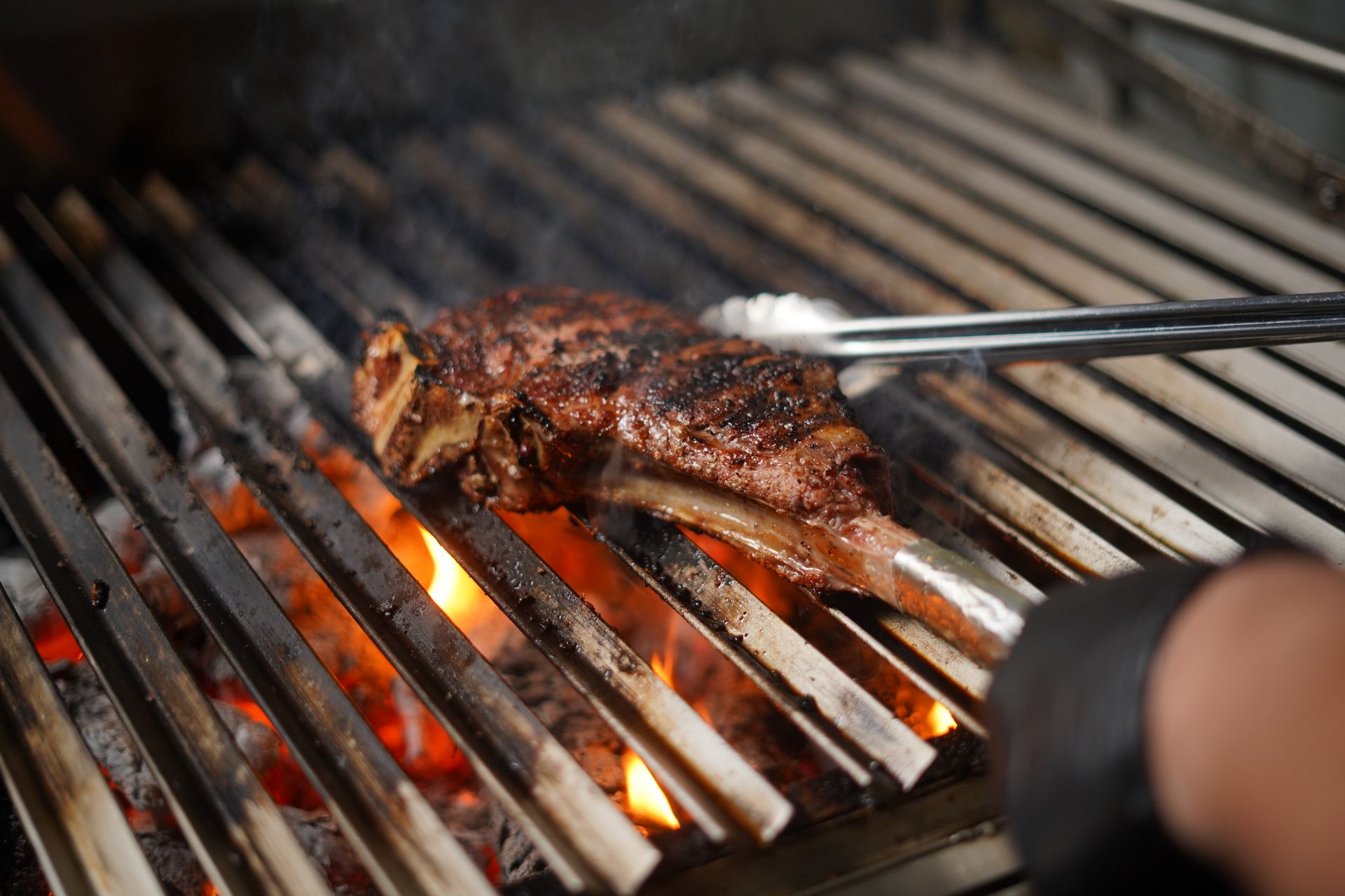Tomahawk steak turned with tongs over an open charcoal grill
