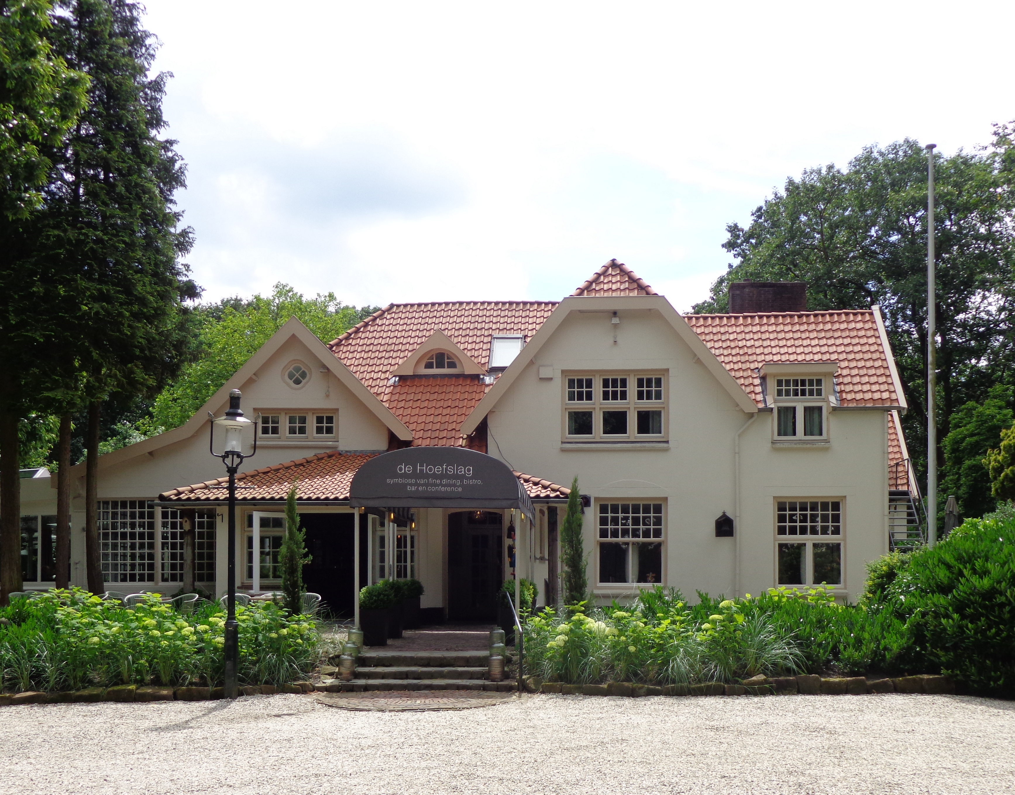 White villa with a red tiled roof and a dark entrance awning among tall trees