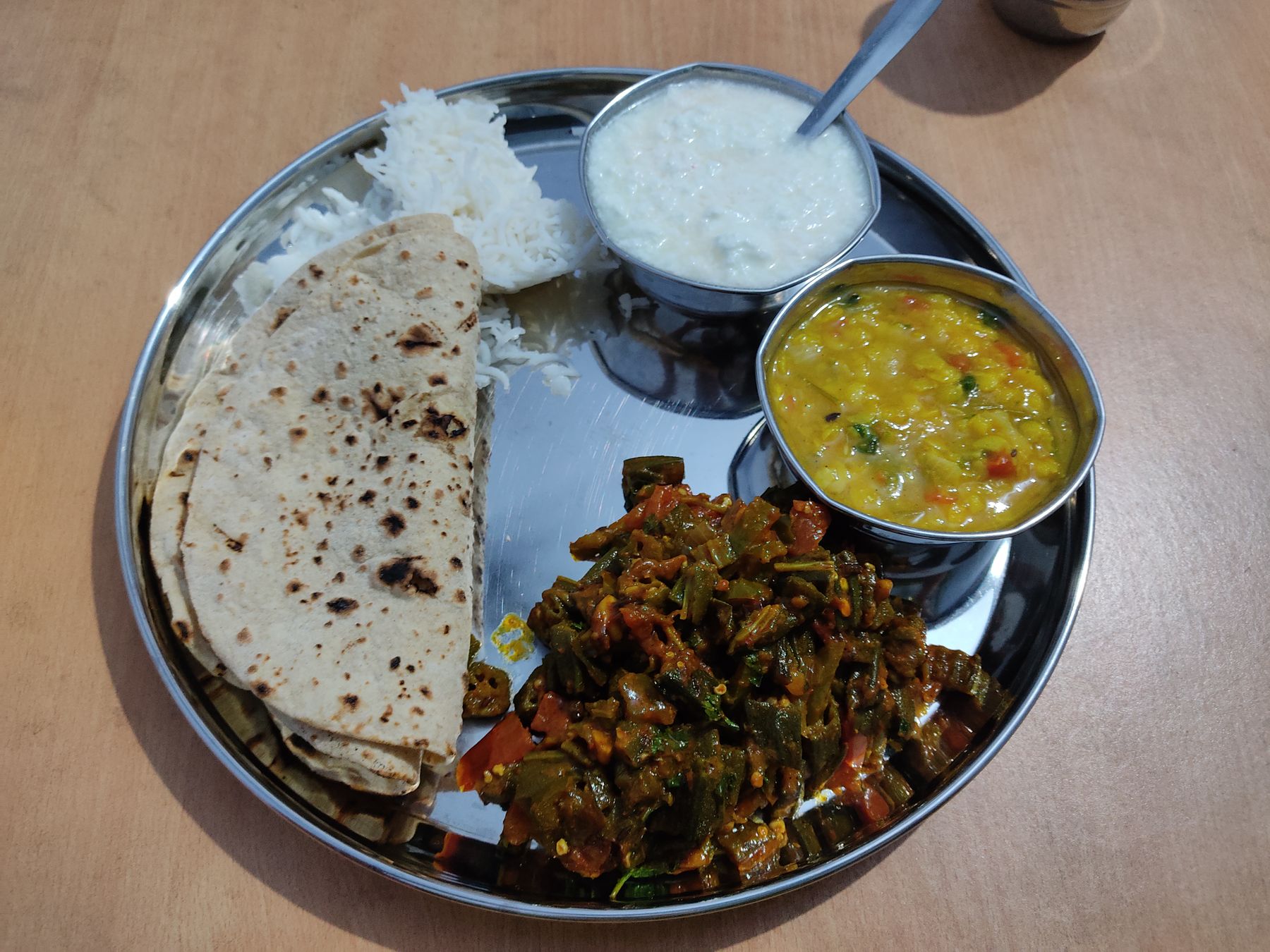 Plato thali de acero con roti, arroz, dal y okra guisada sobre una mesa de madera