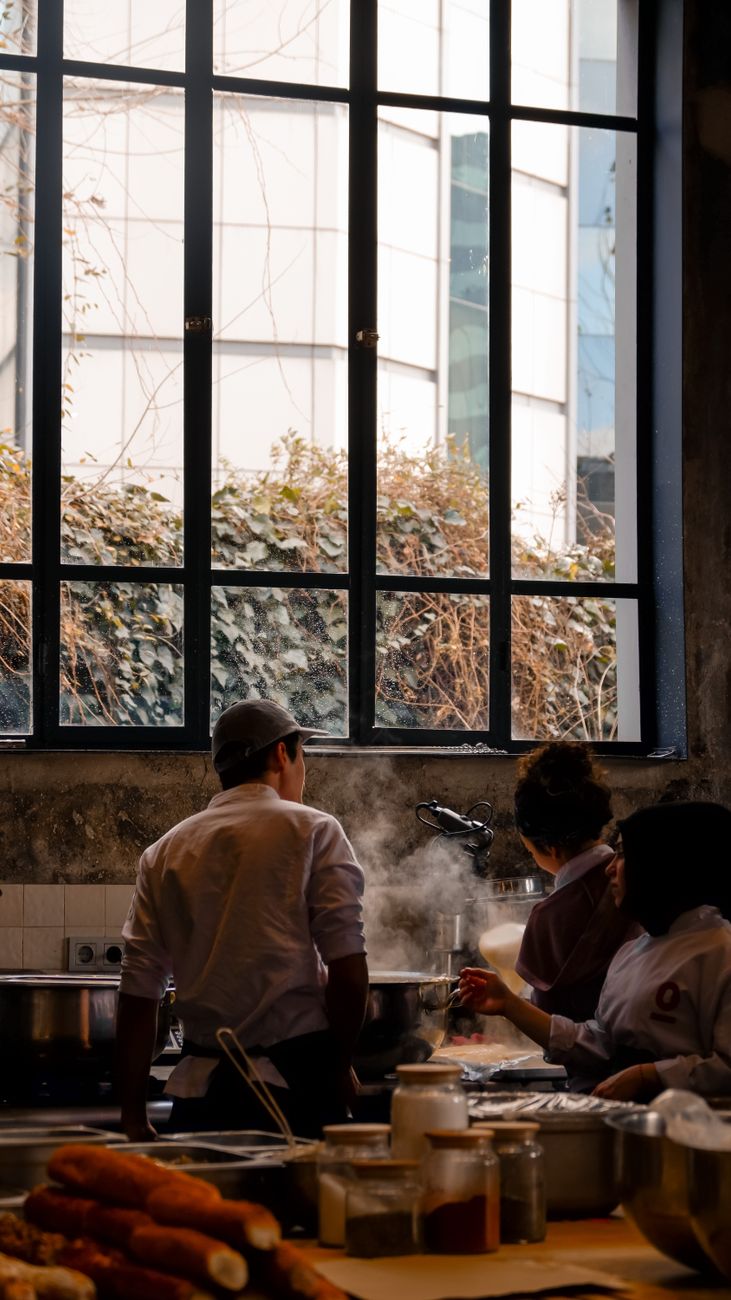 Junge Köche arbeiten nebeneinander an dampfenden Pfannen in einer offenen Restaurantküche