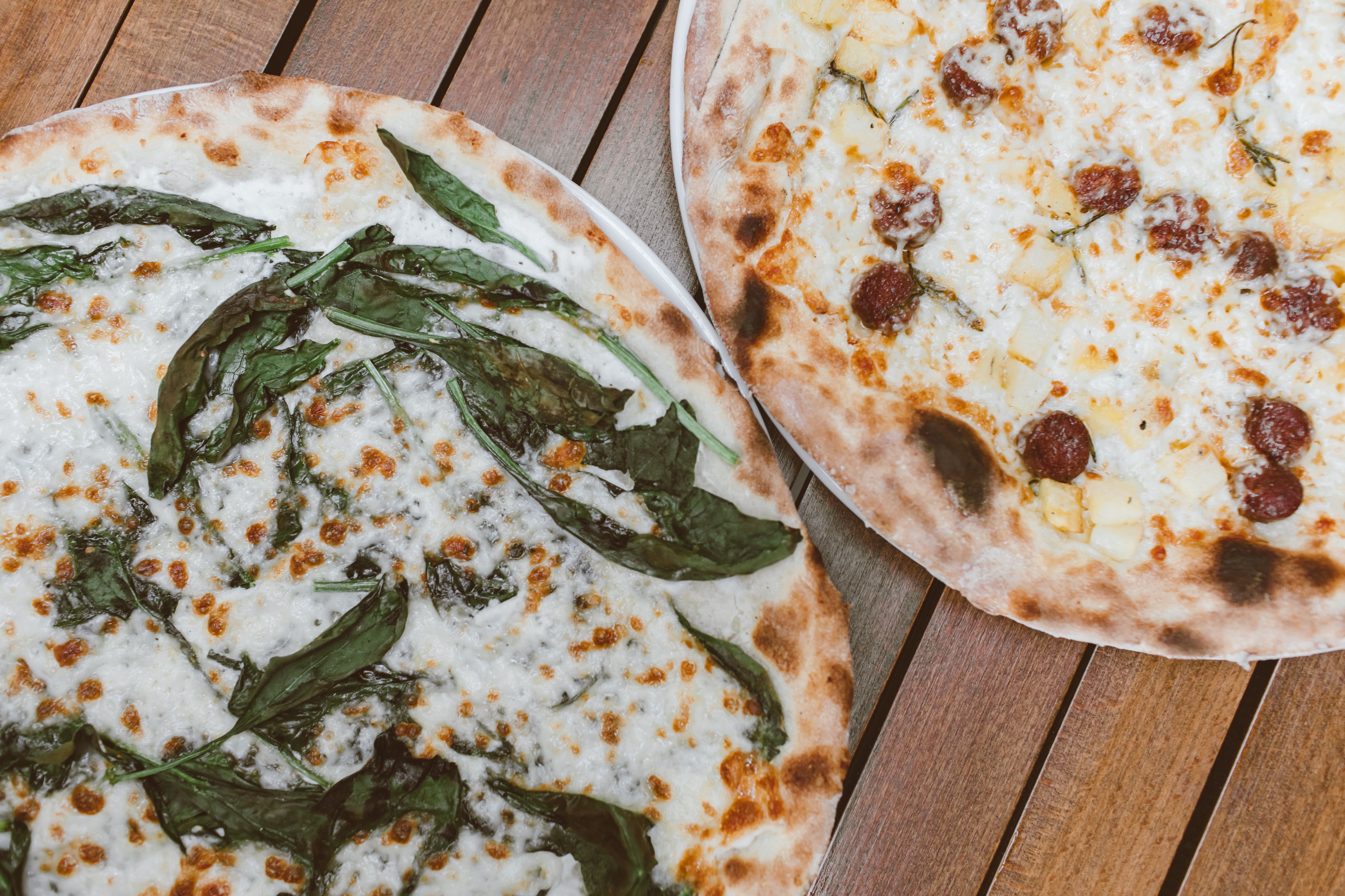 Two round pizzas with spinach and pepperoni on a wooden restaurant table
