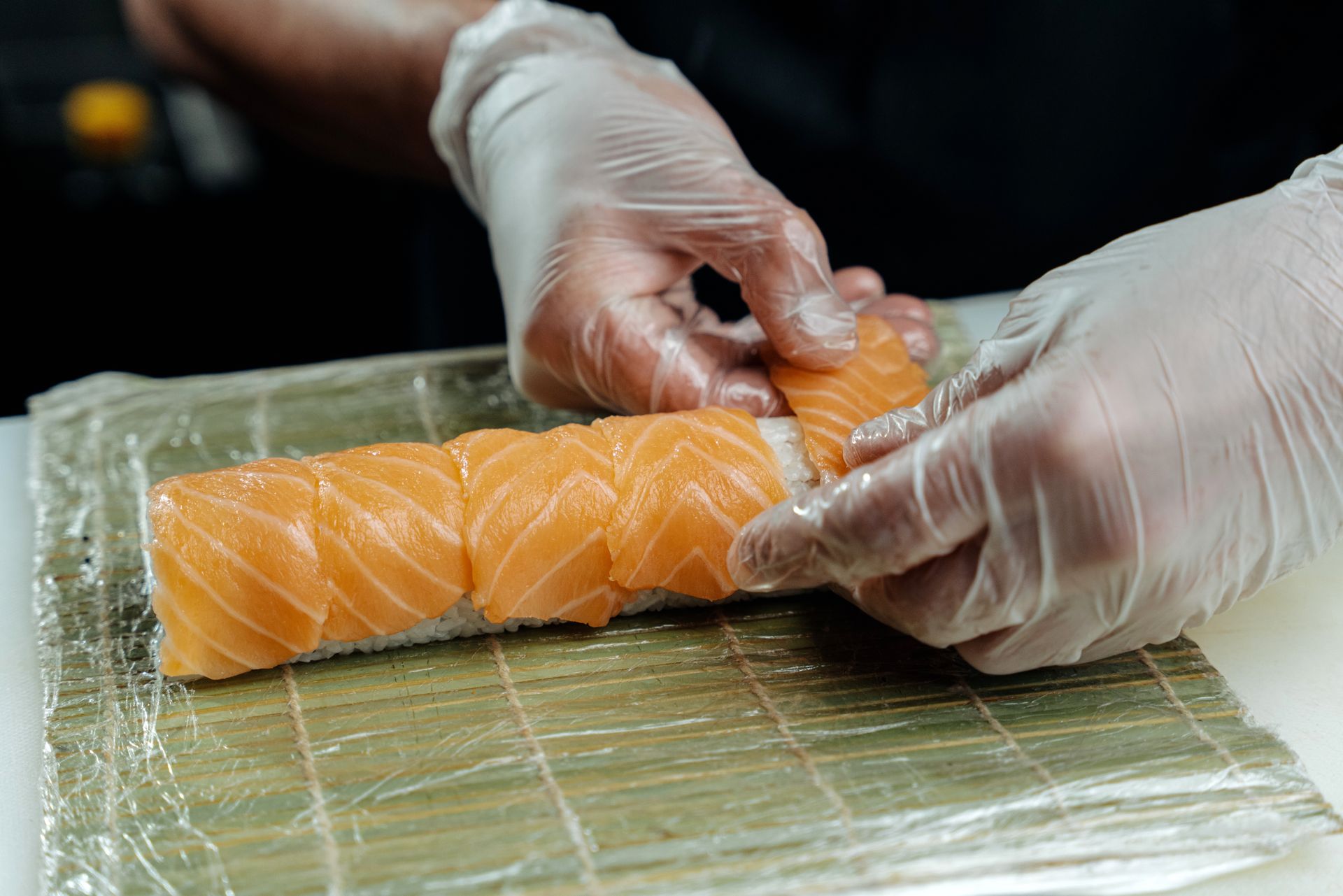 Gloved hands laying slices of salmon over rice on a bamboo mat