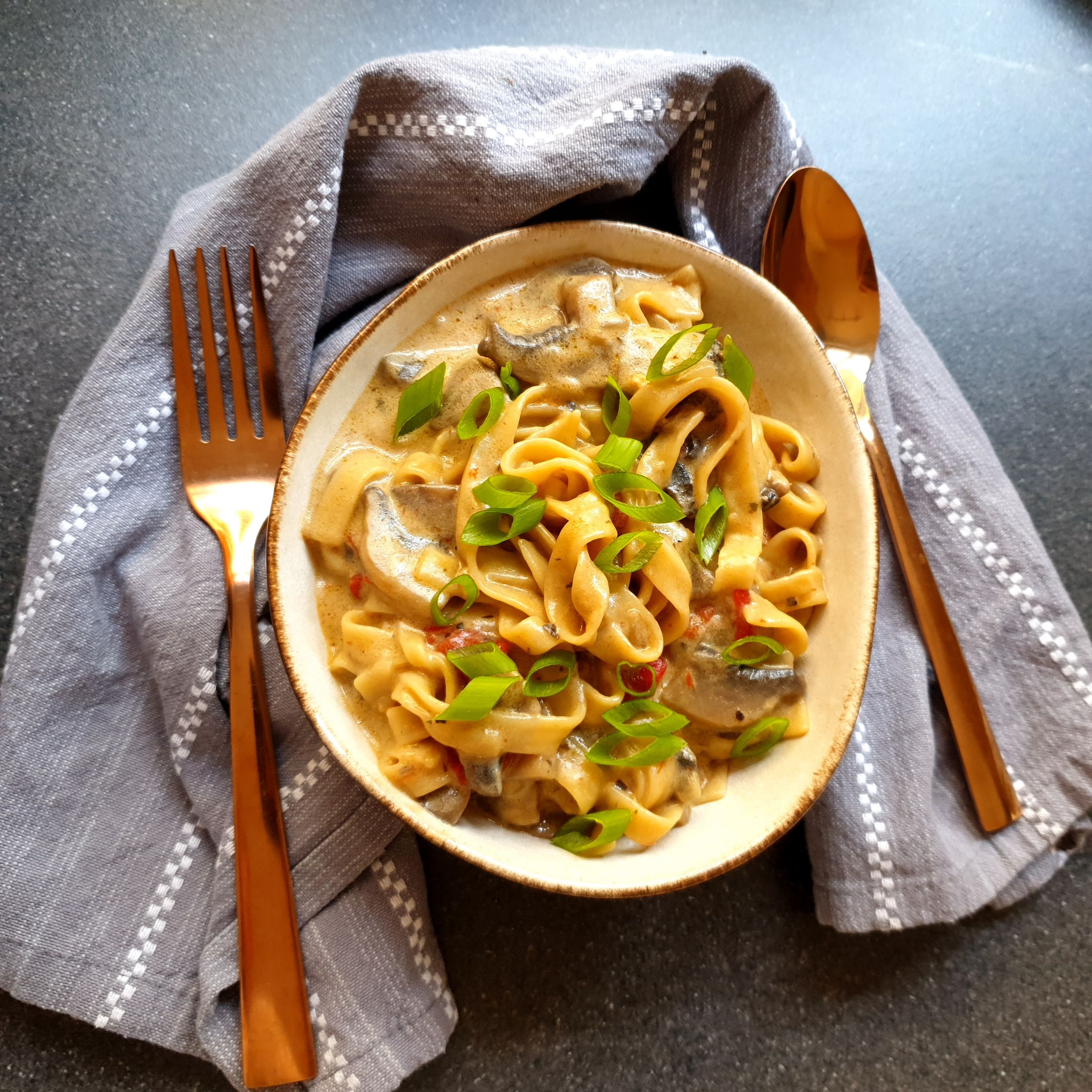 Bowl of tagliatelle pasta with creamy mushroom sauce and spring onion, fork and spoon beside it