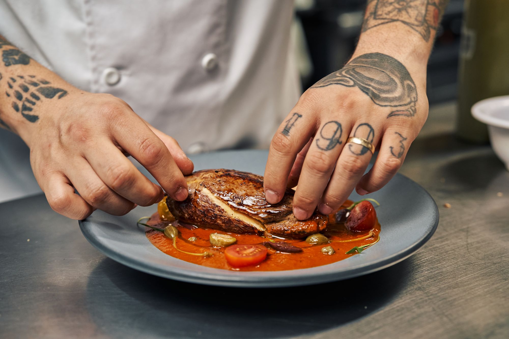 Tattooed chef's hands placing a piece of meat on a grey plate with sauce
