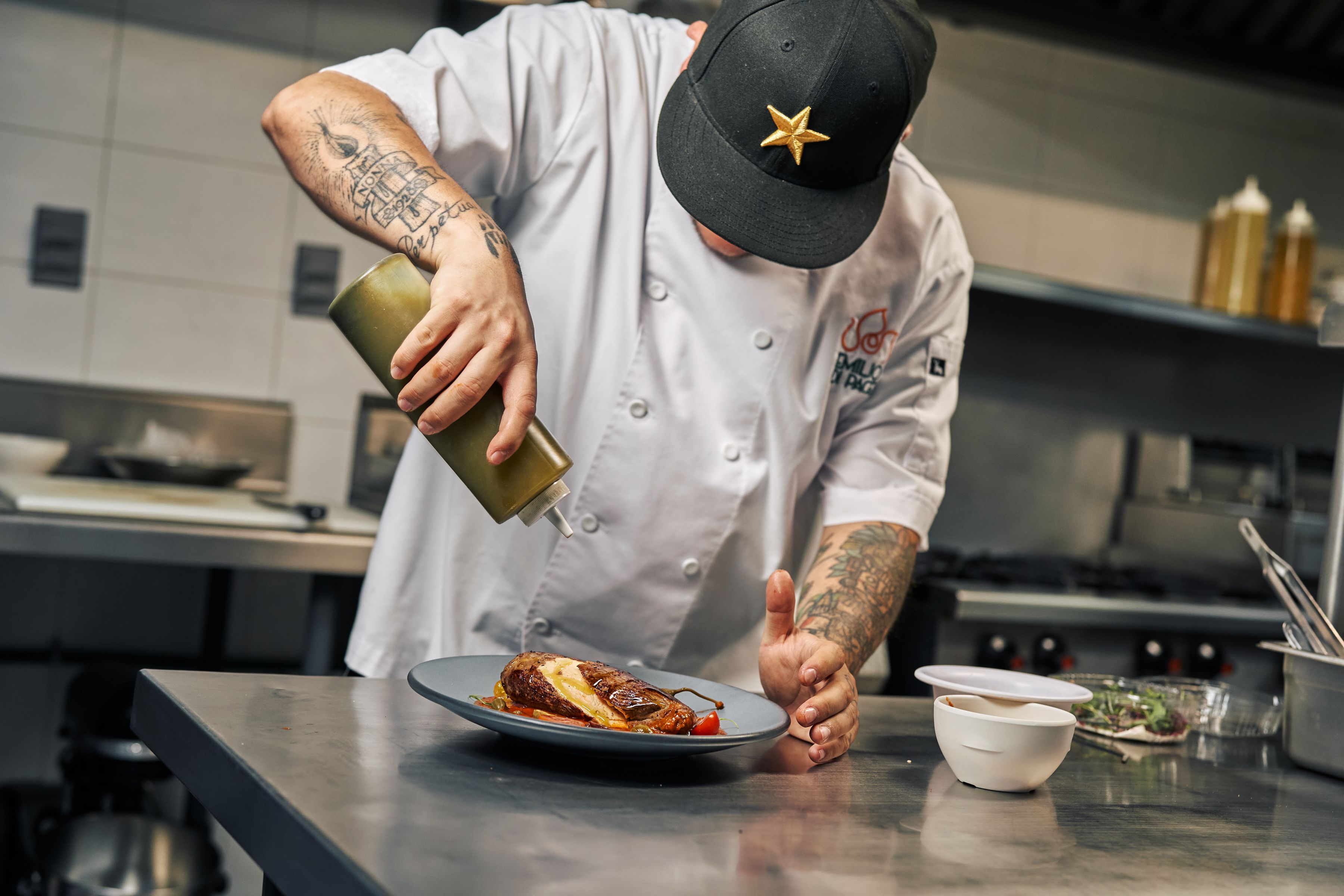 Chef in white jacket and black cap garnishing a plated dish in a professional kitchen