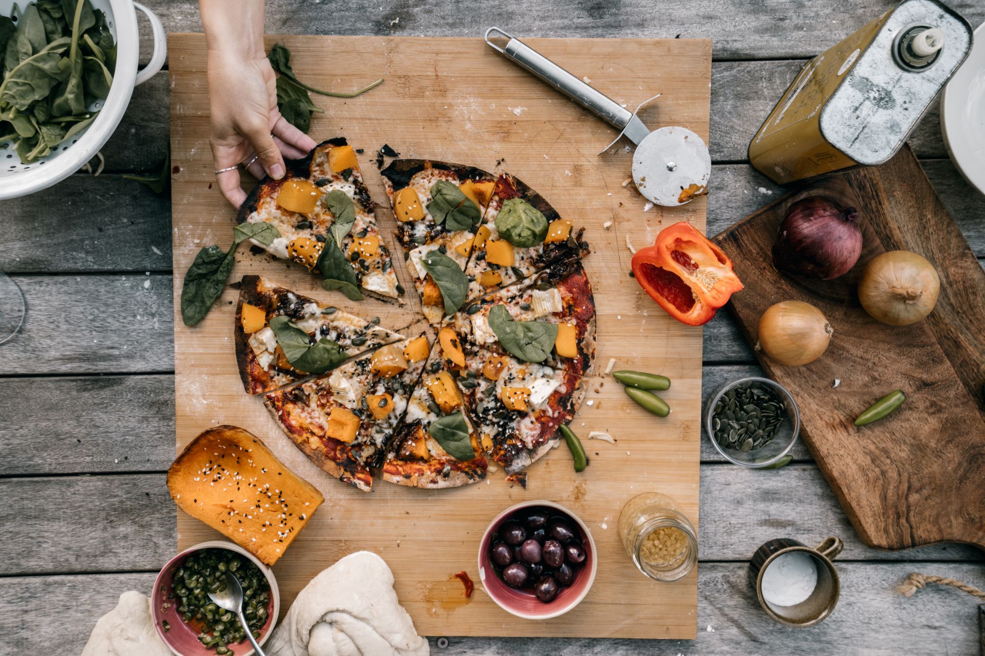 Sliced vegetable pizza on a board surrounded by squash, olives and onions