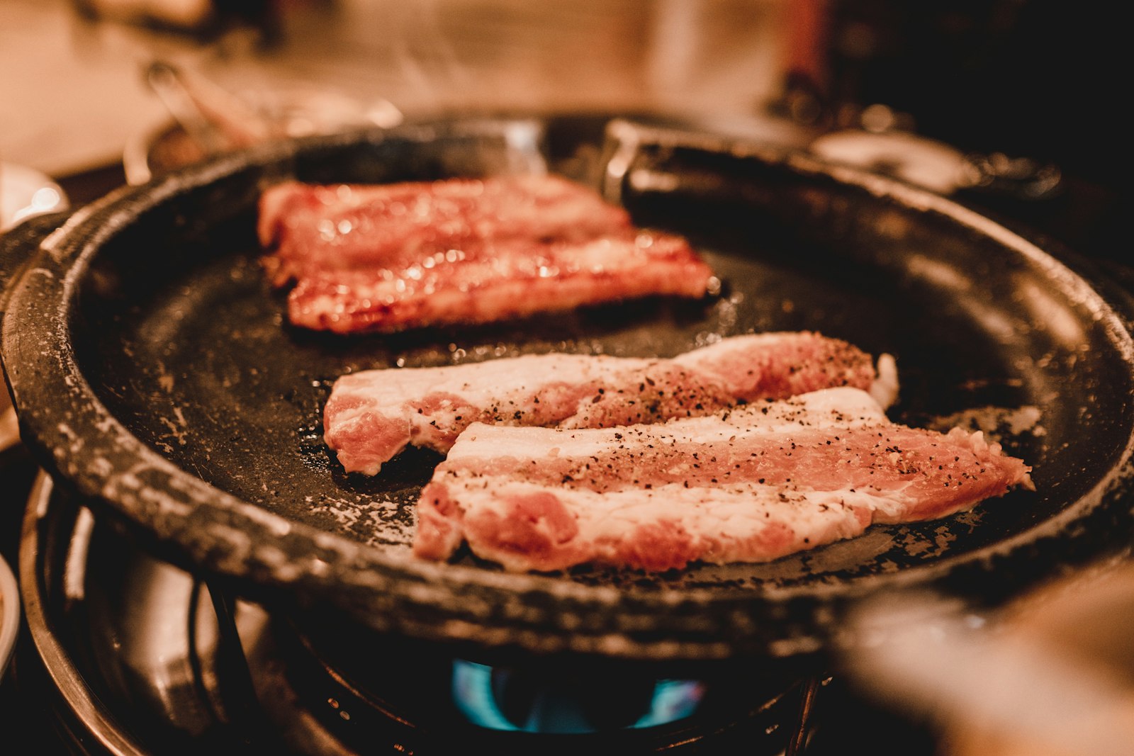 Slices of pork belly seasoned with black pepper grilling on a round cast-iron pan over a gas flame