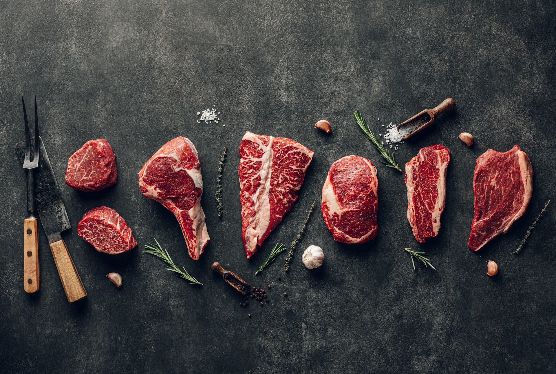 Raw beef steaks of different cuts laid out on a dark stone surface with herbs