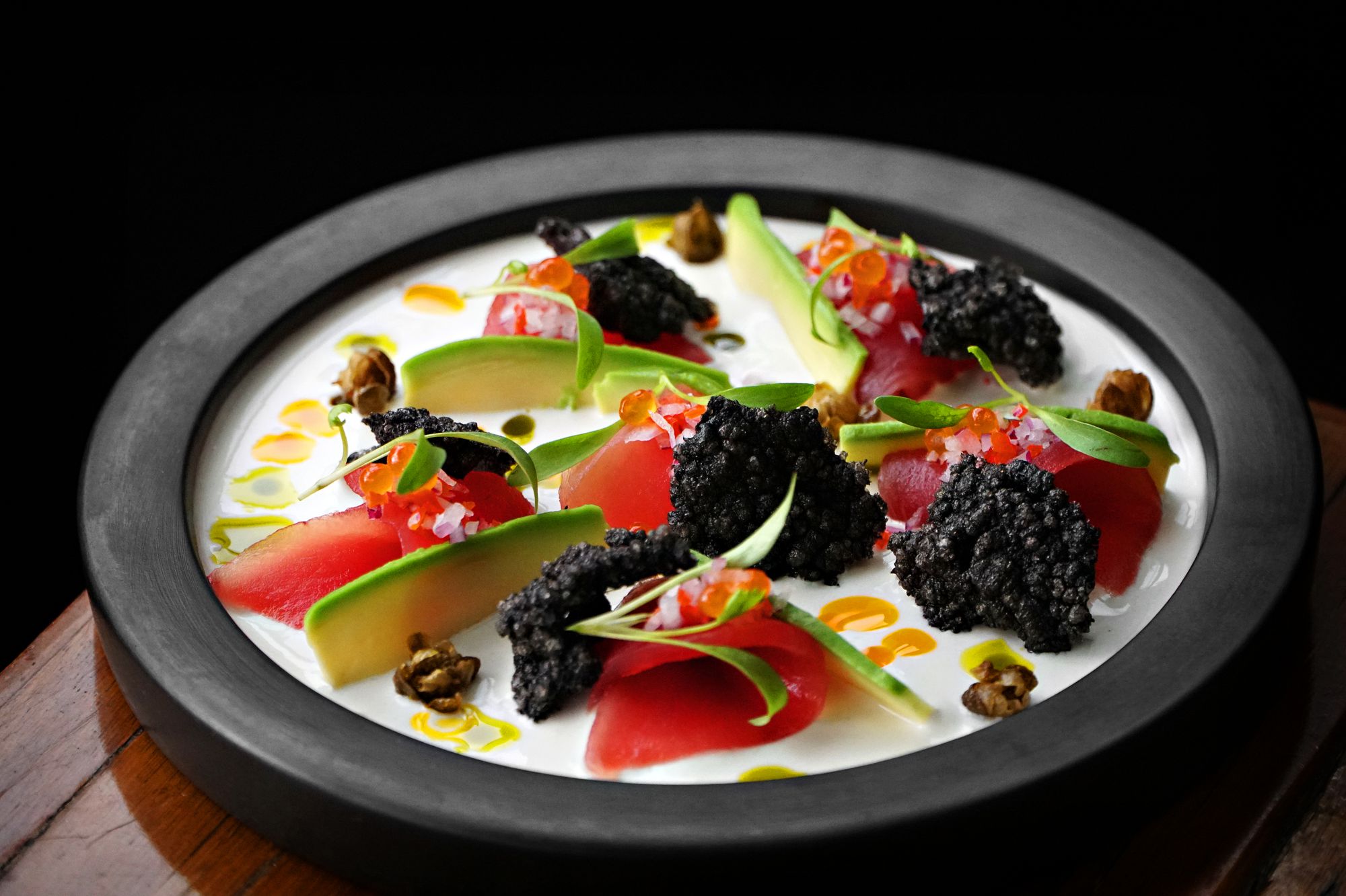 Raw fish slices with avocado, roe and black crisps on a dark ceramic plate