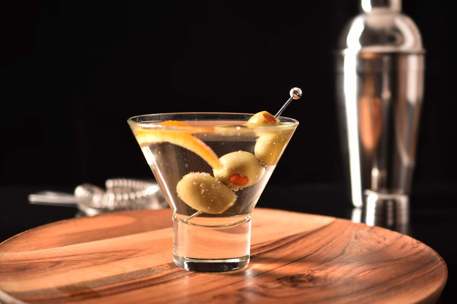 Classic martini with green olives and orange peel on a wooden tray beside a cocktail shaker