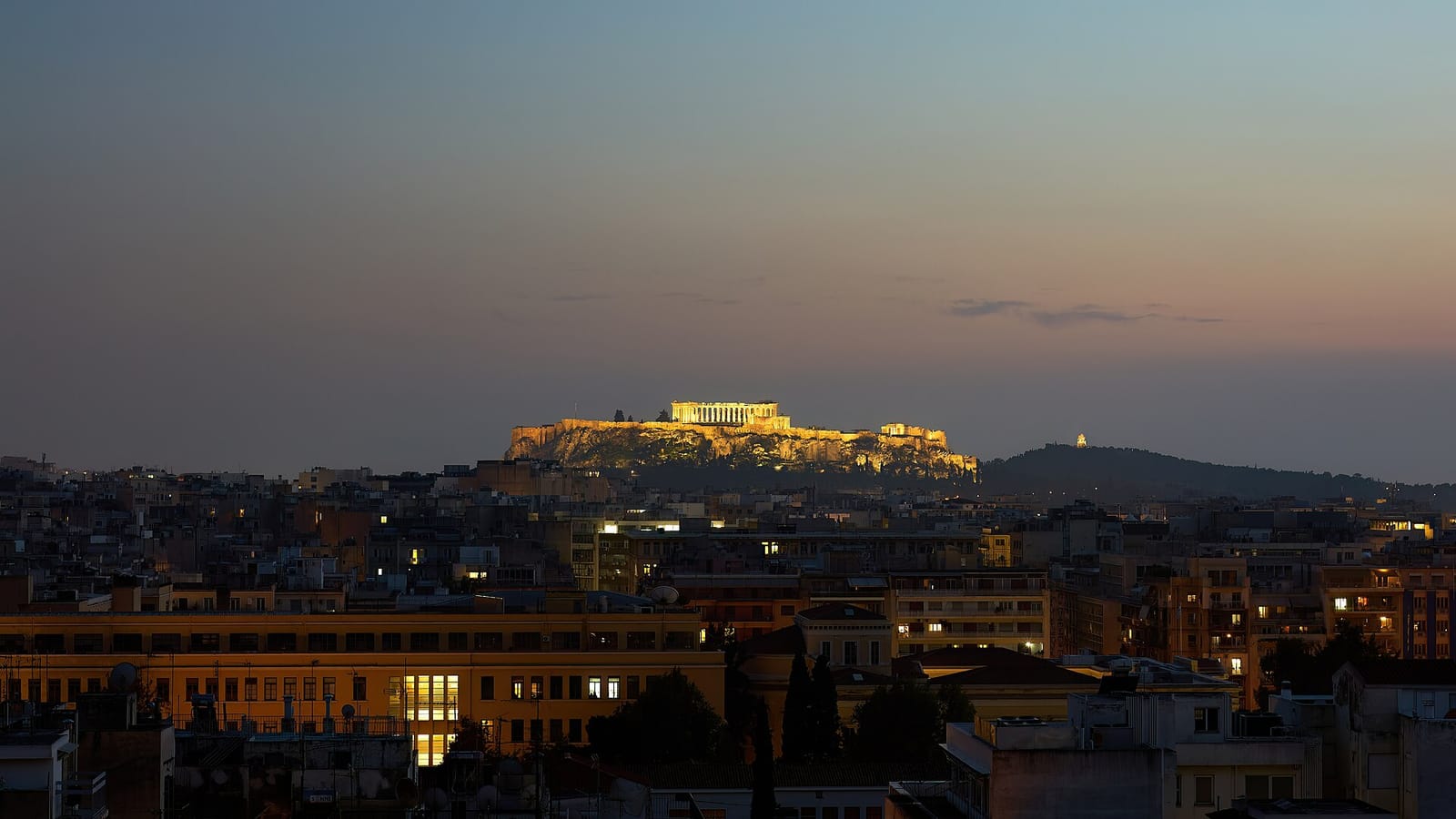 De verlichte Akropolis boven de daken van het centrum van Athene vlak na zonsondergang