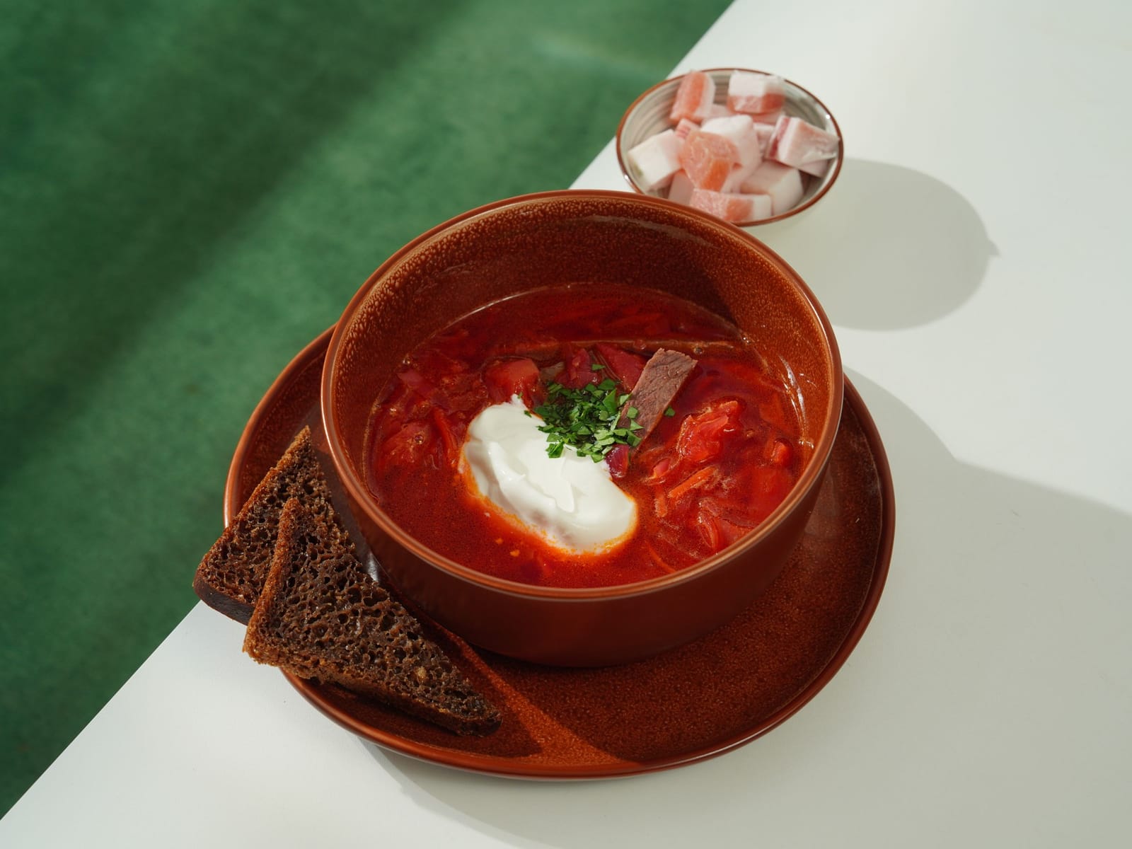 A bowl of red borscht with a spoonful of sour cream and chopped herbs, rye bread and cubes of salo on the side