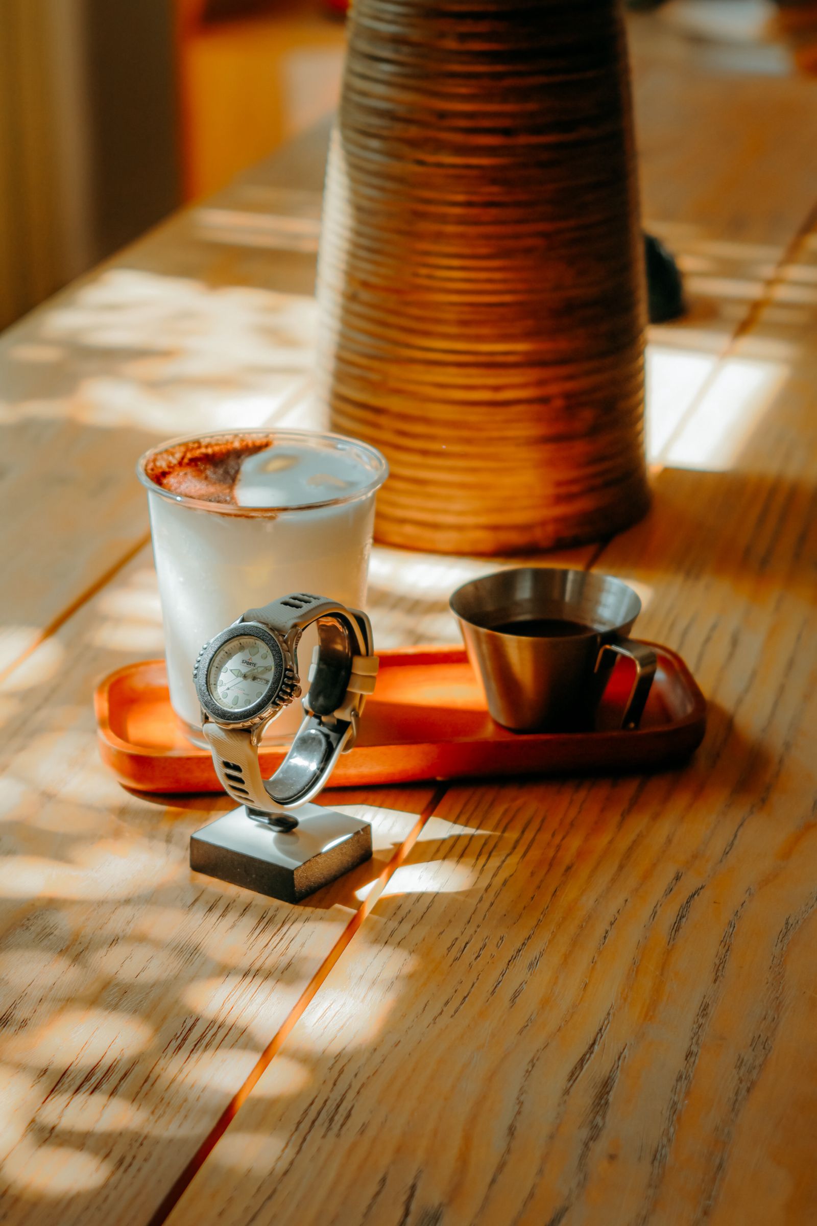 A coffee-themed wristwatch displayed on a wooden counter beside an iced coffee and an espresso cup