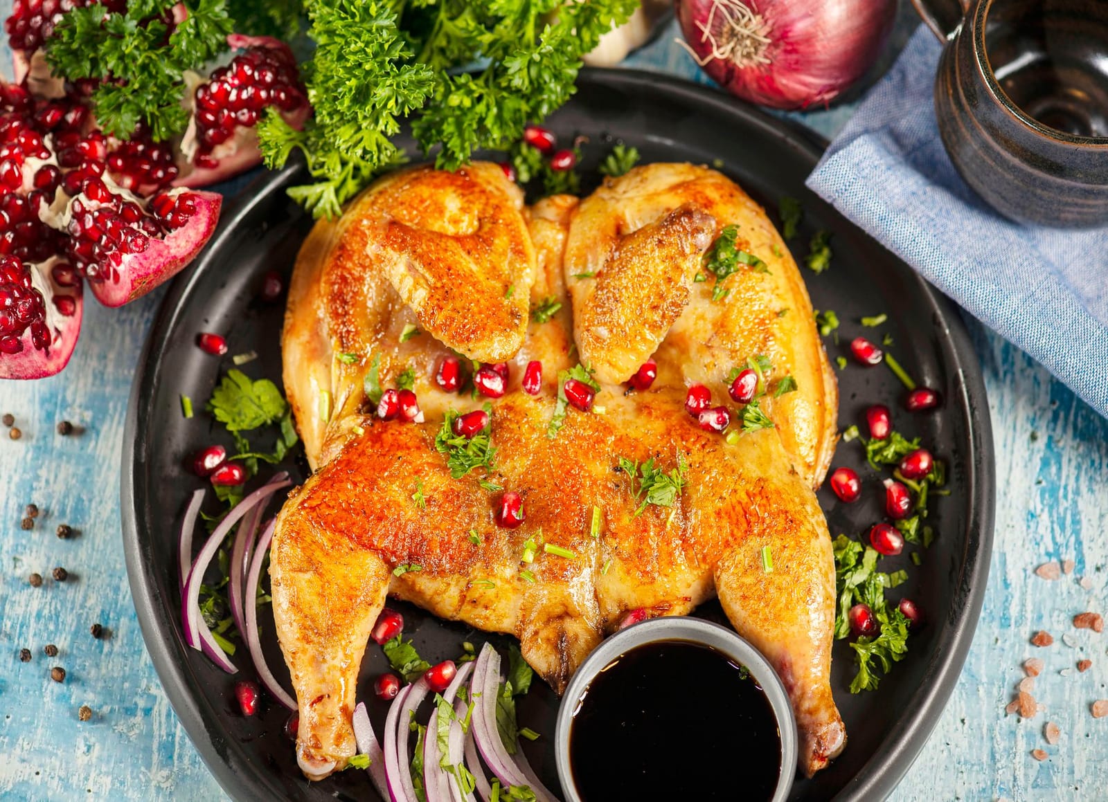 Roasted chicken topped with pomegranate seeds and herbs beside fresh pomegranate halves