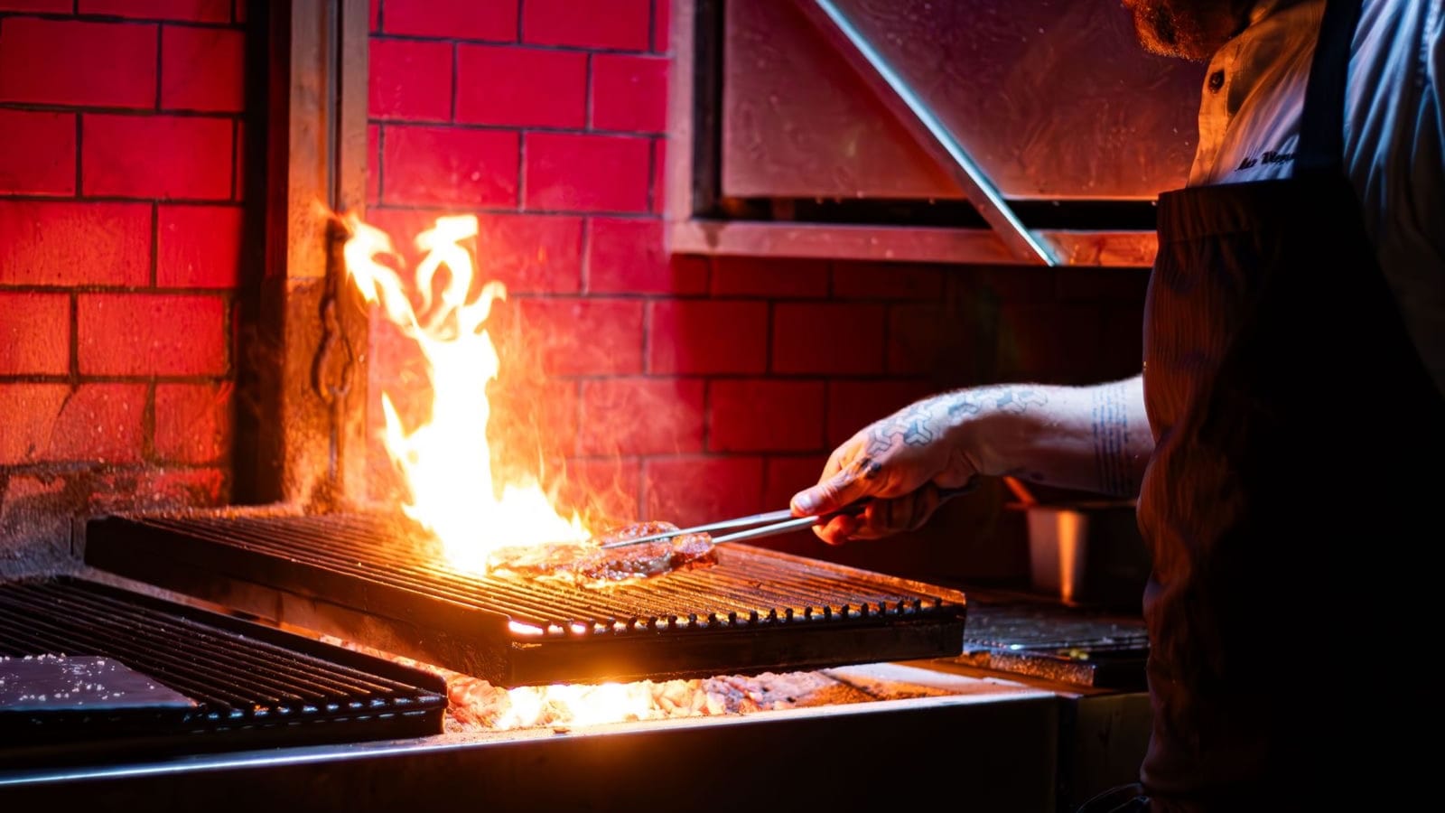A chef tends an open charcoal flame grill in a restaurant kitchen