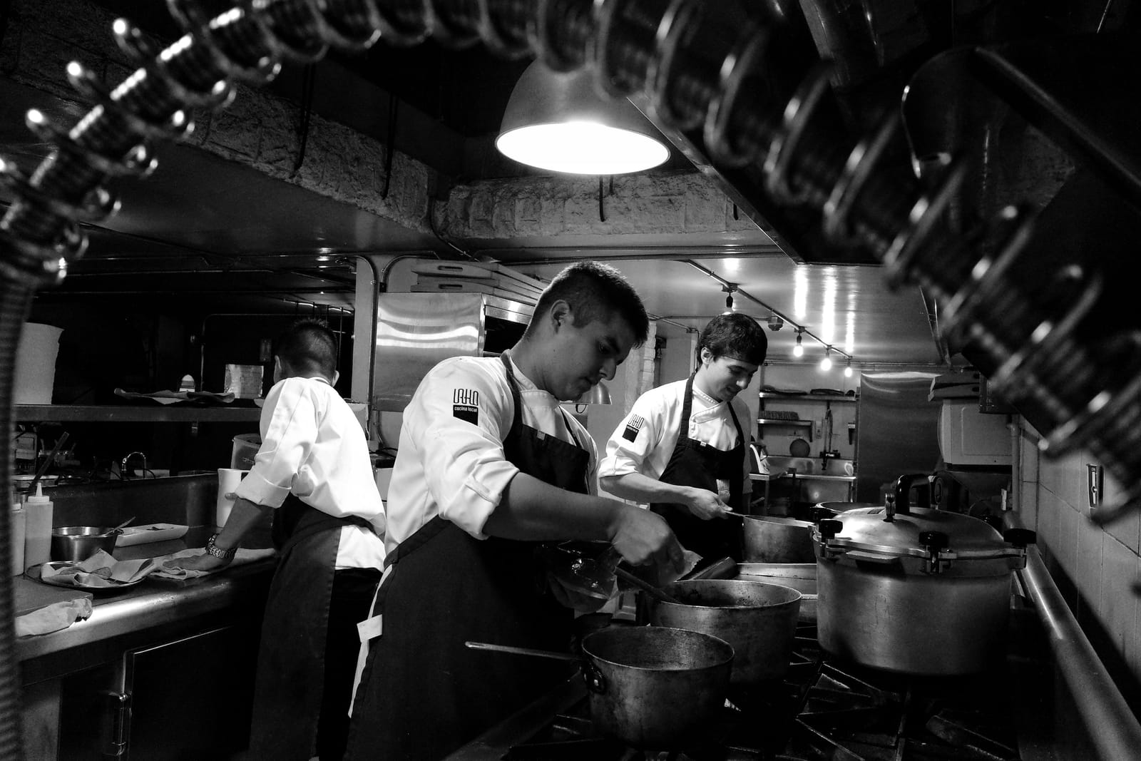 Zwart-witfoto van koks die werken aan het fornuis van een restaurantkeuken onder hanglampen
