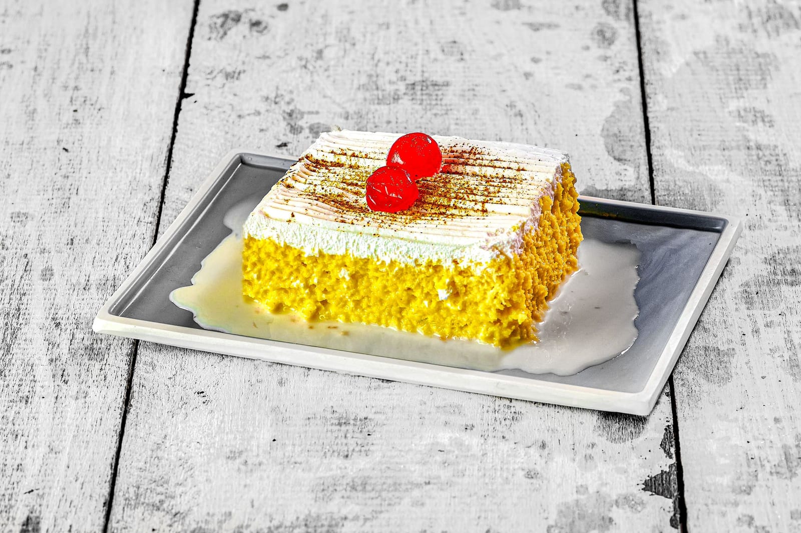 Un quadrato di torta tres leches con panna, cannella e due ciliegie in una pozza di latte