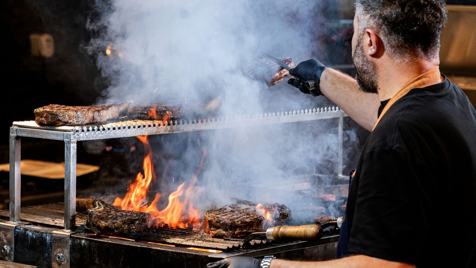 Chef grilling steaks over an open flame on a wood-fired restaurant grill
